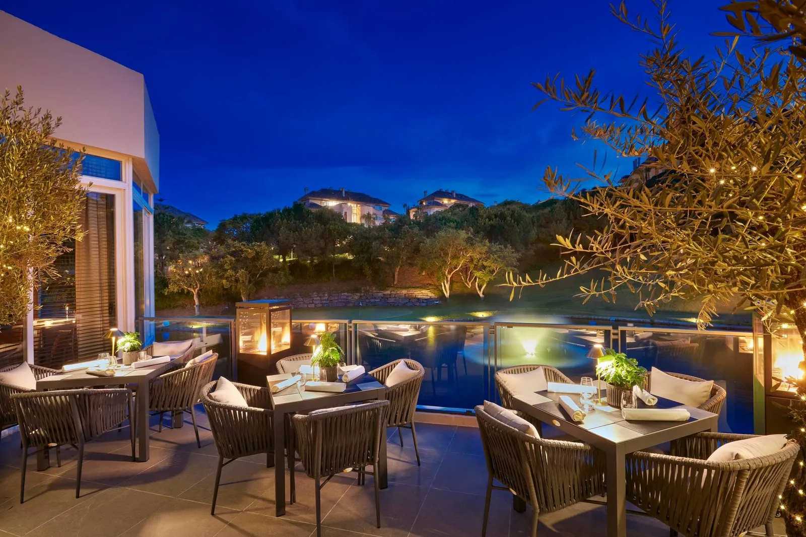 The terrace at dusk, set tables beside the illuminated infinity edge and the lake