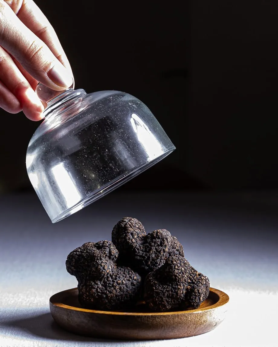 A hand lifting a glass cloche to reveal fresh black truffles on a bronze dish