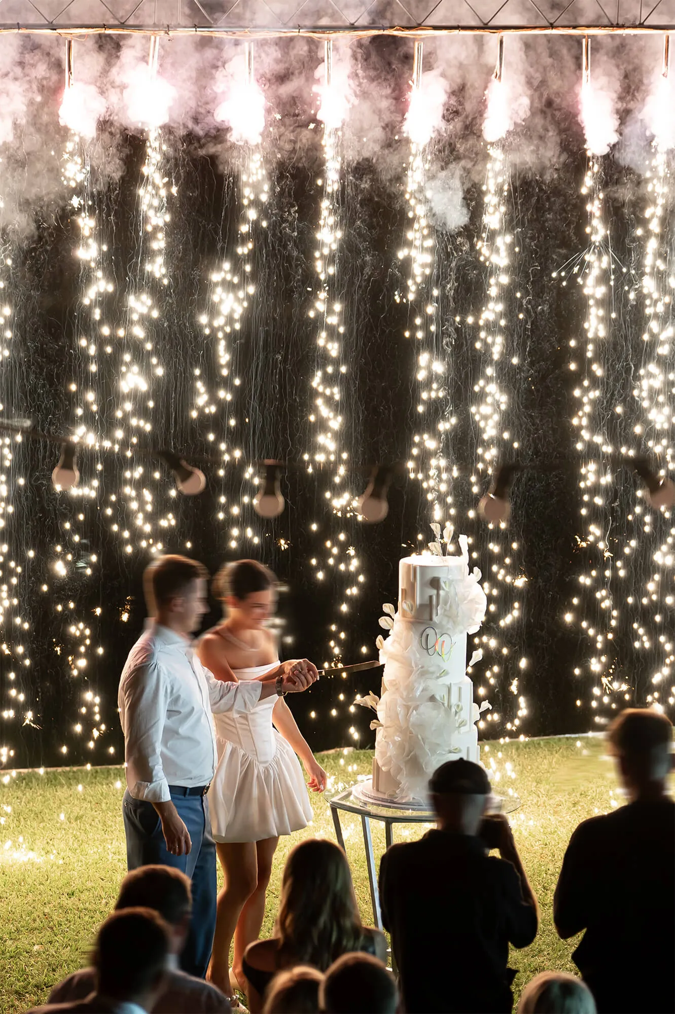 A couple cutting their wedding cake at night under a curtain of lights and sparklers